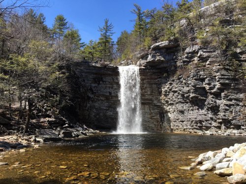 A serene waterfall cascades into a clear pool, surrounded by rocky cliffs and lush trees under a blue sky.