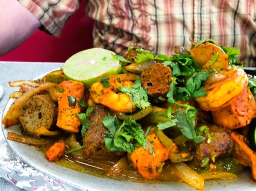 A colorful platter of shrimp and meat skewers, garnished with cilantro and lime, served with sautéed onions and peppers.