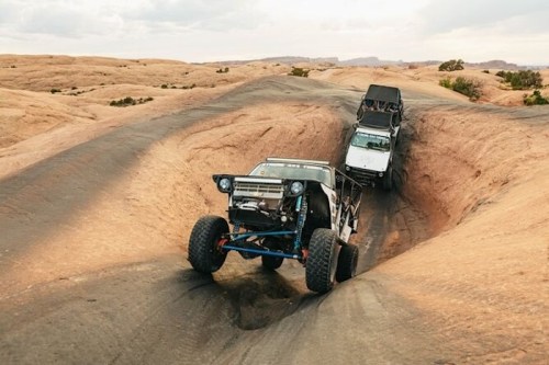 Two off-road vehicles navigate a rugged, rocky terrain in a desert landscape.