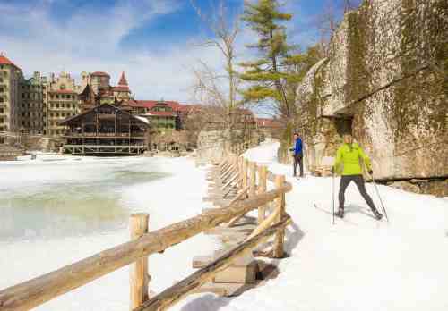A person skiing on a snowy path beside a frozen lake, with historic buildings and trees in the background.