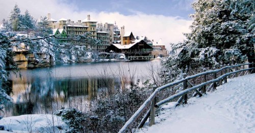 A snowy landscape with a lake and rustic buildings in the background, surrounded by trees and a wooden path.