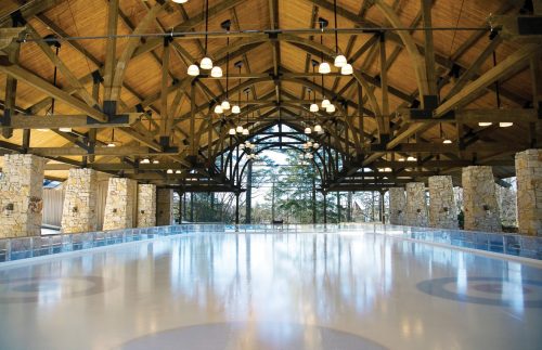 A spacious indoor ice rink with wooden beams and stone walls, illuminated by hanging lights.