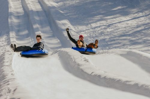 Two people joyfully tubing down a snowy slope, with bright blue tubes and a clear winter sky in the background.