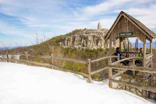 Two hikers stand on a wooden platform overlooking a snowy landscape and a rocky hill with a tower in the distance.