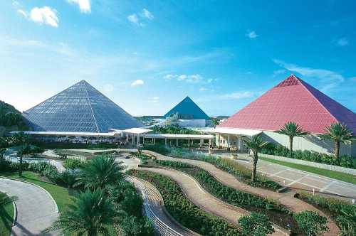 Three large pyramids with glass and colored roofs, surrounded by lush greenery and a clear blue sky.