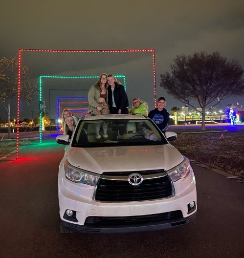 A group of friends poses on a car in front of colorful light arches at night.
