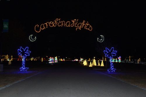 A festive entrance archway reading "Carolina Lights" with colorful holiday lights lining the pathway and trees.