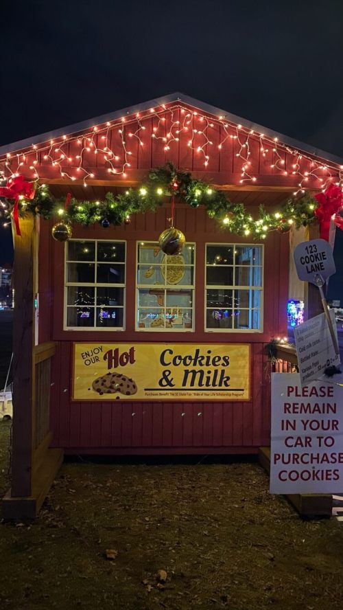 A festive cookie stand with holiday lights, a sign for hot cookies and milk, and a message to remain in your car for purchases.