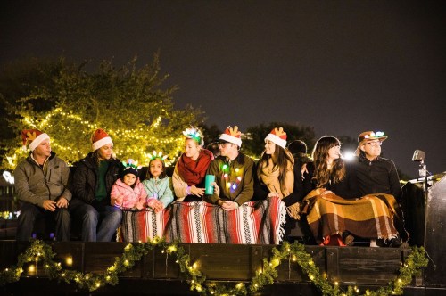 A group of people wearing festive hats and blankets sit together, surrounded by holiday lights at night.