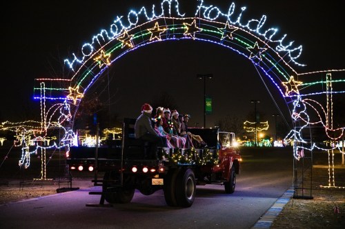 A decorated truck drives under a colorful light arch reading "Hometown Holidays" at night.