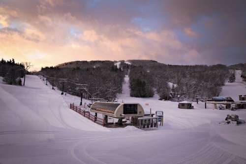 A snowy ski resort with a lift station, surrounded by trees and a colorful sunset in the background.