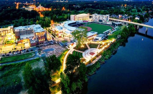 Aerial view of a riverside park and sports complex, with buildings and lights illuminating the area at dusk.