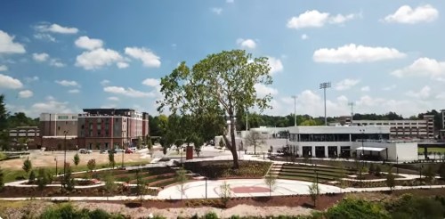 A sunny view of a park with a large tree, surrounded by buildings and a sports field in the background.