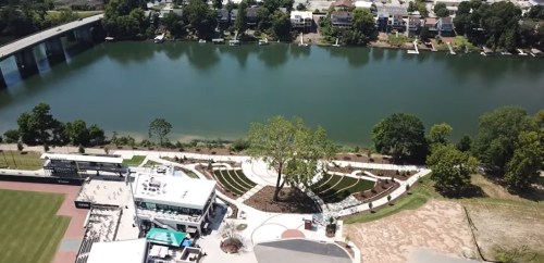 Aerial view of a park by a river, featuring green spaces, pathways, and nearby buildings along the water's edge.