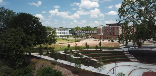 Aerial view of a park with green spaces, pathways, and modern buildings under a partly cloudy sky.