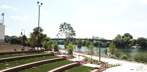 A scenic view of a park with green lawns, steps, and a river under a clear sky, featuring a bridge in the background.