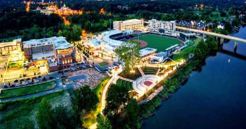 Aerial view of a vibrant riverside area featuring a baseball field, park, and illuminated buildings at dusk.