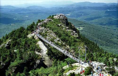 A suspension bridge connects rocky peaks, surrounded by lush green forests and distant mountains under a clear blue sky.