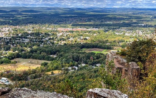 A panoramic view of a lush valley with fields, trees, and a small town under a partly cloudy sky.