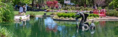 A serene park scene featuring a pond, colorful flowers, a red bridge, and people enjoying the outdoors.