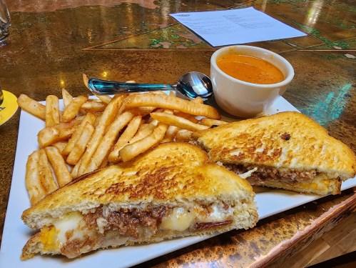 A plate with a grilled sandwich filled with meat and cheese, served with crispy fries and a bowl of soup.