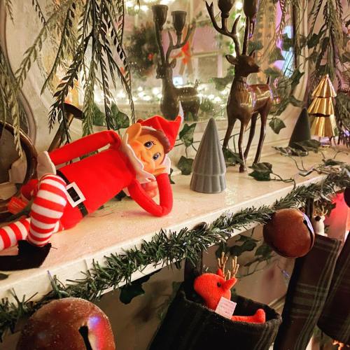 A festive shelf decorated with greenery, candles, and holiday ornaments, featuring a playful elf doll lounging.