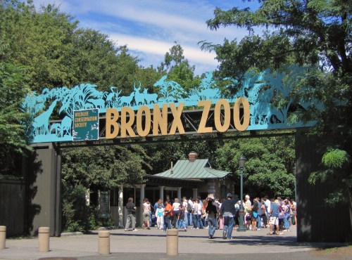 Entrance to the Bronx Zoo, featuring a large sign and a crowd of visitors under a blue sky.