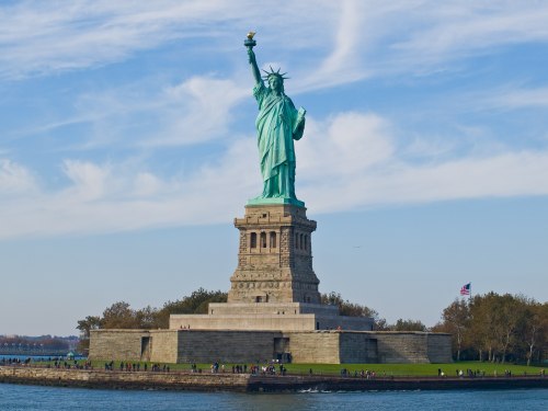 The Statue of Liberty stands tall on Liberty Island, with a clear blue sky and people gathered around its base.