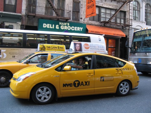 A yellow NYC taxi drives past a deli and grocery store on a busy street. Buses and other vehicles are in the background.