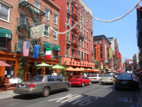 A vibrant street scene with red brick buildings, outdoor dining, flags, and cars parked along the road in a lively neighborhood.