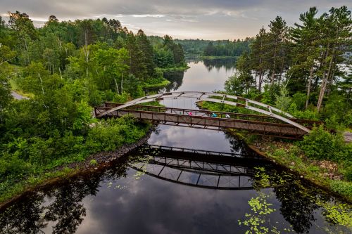 Aerial view of a bridge over a calm lake, surrounded by lush greenery and trees under a cloudy sky.