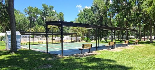 A shaded outdoor pickleball court with benches, surrounded by trees and blue skies.