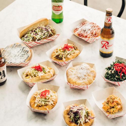 A variety of colorful dishes in paper trays, including hot dogs, tacos, and sides, with drinks on a table.