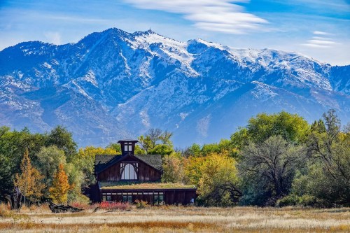 A rustic wooden building surrounded by colorful trees, with snow-capped mountains in the background under a blue sky.