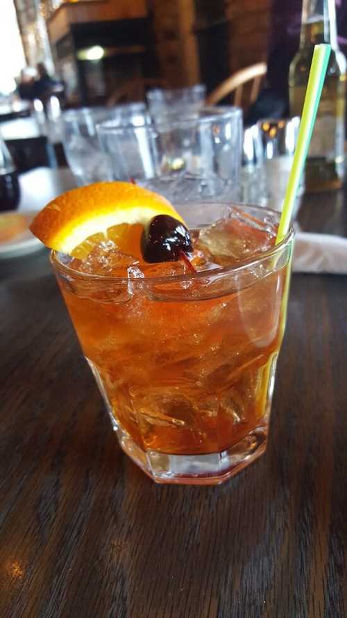 A cocktail in a glass with ice, garnished with an orange slice and a cherry, on a wooden table.