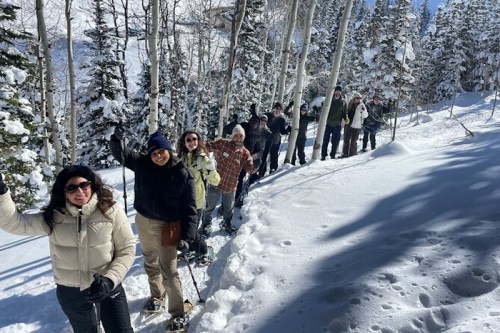 A group of people snowshoeing through a snowy forest, smiling and waving at the camera.