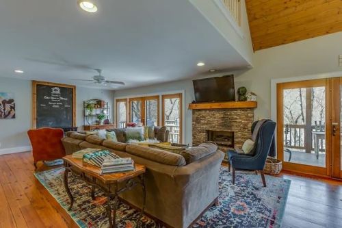 Cozy living room with a stone fireplace, large windows, and comfortable seating, featuring wooden accents and a colorful rug.