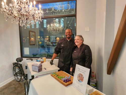 Two smiling chefs stand behind a table with colorful desserts, set in a bright shop with a chandelier in the background.