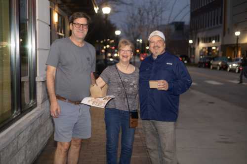 Three smiling people stand on a sidewalk at dusk, holding takeout boxes and enjoying a night out.