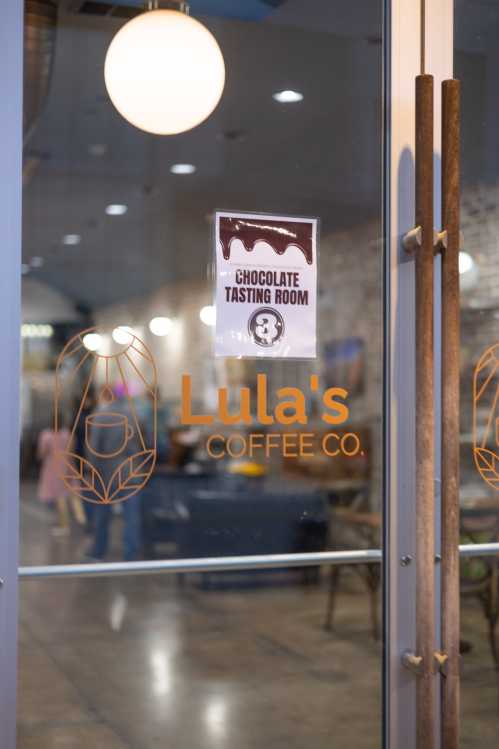 Sign on a glass door reads "Chocolate Tasting Room" at Lula's Coffee Co., with a cozy café interior visible.