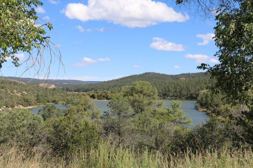 A serene landscape featuring a lake surrounded by trees and rolling hills under a blue sky with fluffy clouds.