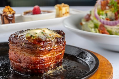 A succulent bacon-wrapped filet mignon on a sizzling plate, with gourmet desserts and a salad in the background.