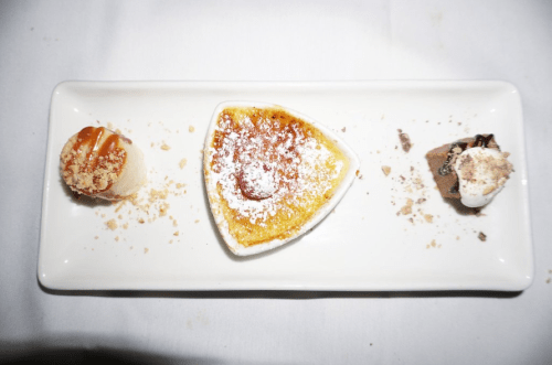 A dessert plate featuring a crème brûlée, a small cake with caramel, and a chocolate mousse, garnished with crumbs.