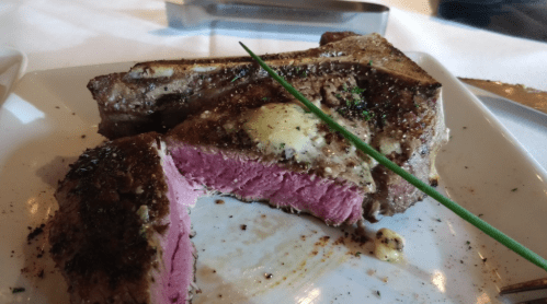 A close-up of a perfectly cooked steak with a pink center, garnished with herbs and a pat of butter on a white plate.