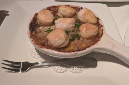 A dish of scalloped seafood topped with golden pastry, served in a white bowl on a lace doily, with a fork beside it.