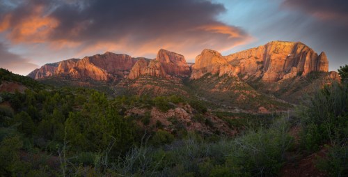 A panoramic view of rugged mountains at sunset, with vibrant colors illuminating the rocky cliffs and surrounding greenery.