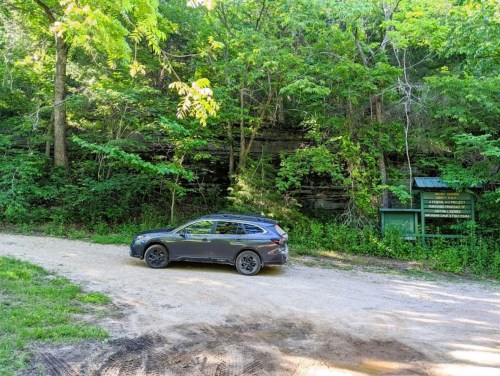 A black SUV parked on a gravel road surrounded by lush green trees and a sign in the background.