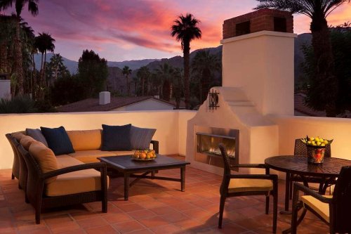 Cozy outdoor patio with a sofa, table, and fireplace, set against a vibrant sunset and palm trees.