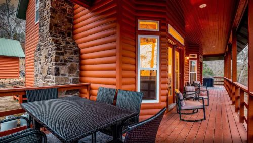 A cozy wooden cabin porch with a stone chimney, featuring a table and chairs, surrounded by nature.