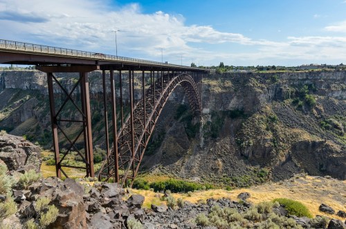 A large metal arch bridge spans a deep canyon, surrounded by rocky terrain and greenery under a blue sky.